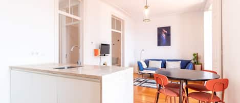 Une cuisine moderne dotée d’un grand îlot central, un coin repas avec une table ronde et des chaises orange, et un salon avec un canapé et un téléviseur.