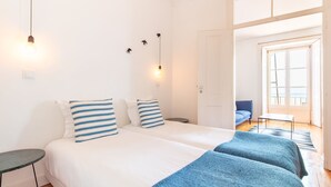 Un lit double avec des oreillers à rayures, une table de chevet, une lampe et une vue sur un canapé bleu et une fenêtre.