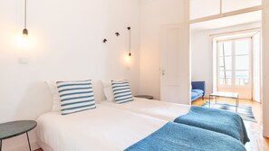 A double bed with striped pillows, a bedside table, a lamp, and a view of a blue sofa and a window. - Alfama Balcony River View by LovelyStay (Lisboa)