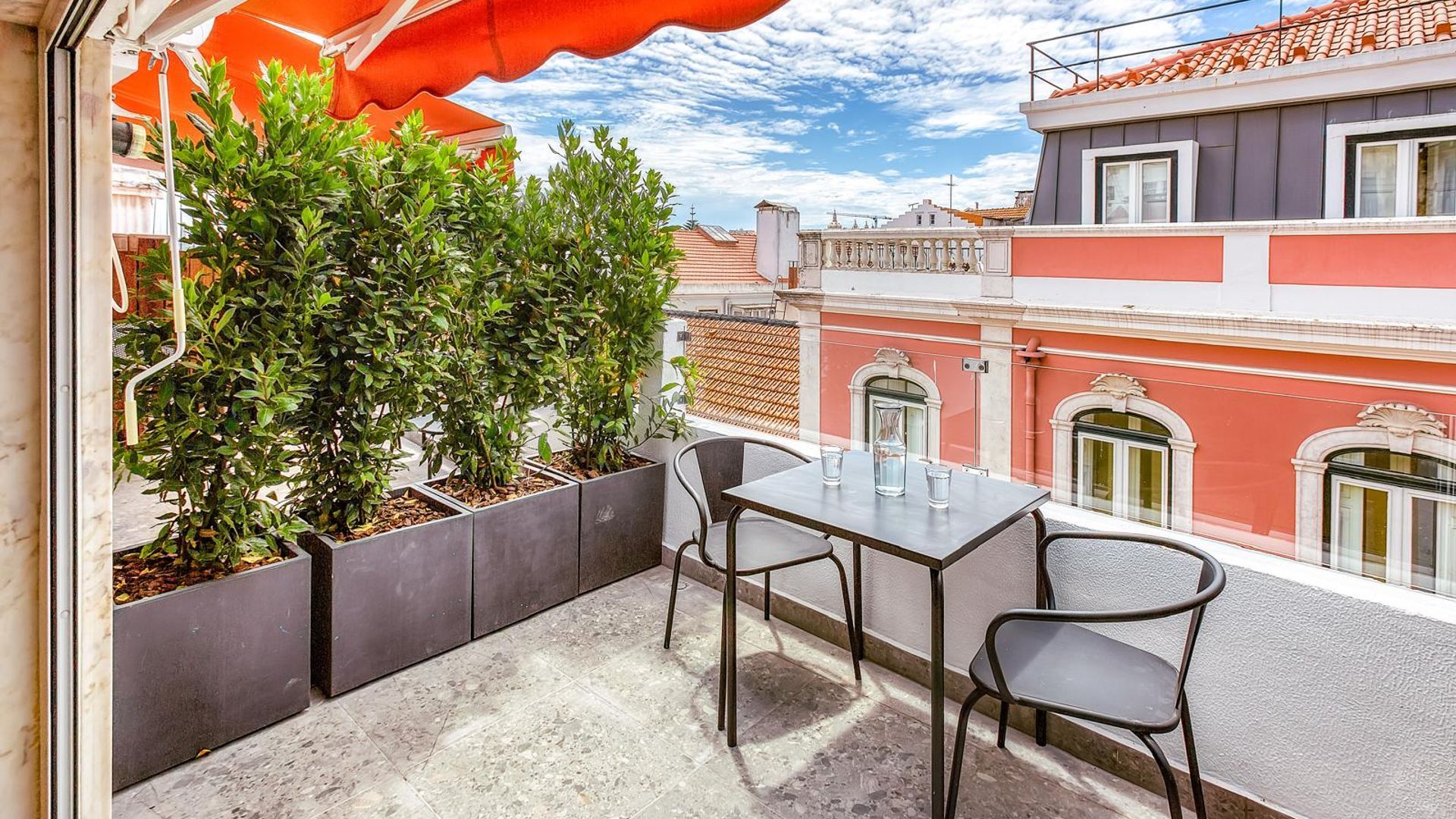 An outdoor terrace with a table and chairs, surrounded by greenery and overlooking a cityscape.