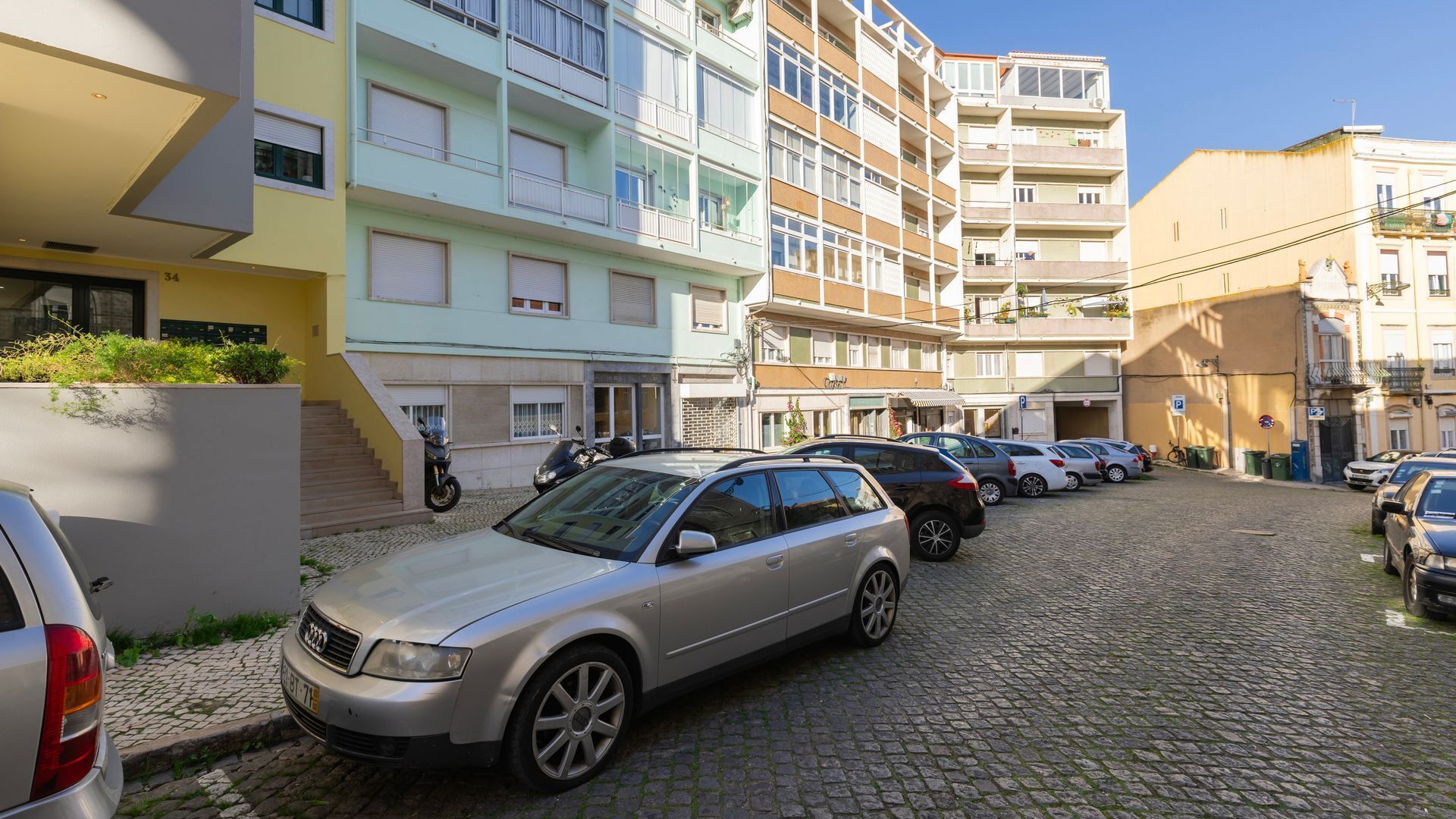 A row of parked cars on a cobblestone street, with multi-story buildings in the background.