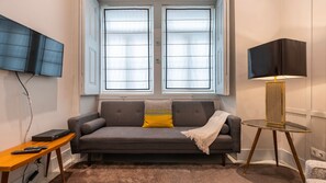 Ein Wohnzimmer mit grauem Sofa, einem kleinen Tisch mit Lampe und einem an der Wand montierten Fernseher.