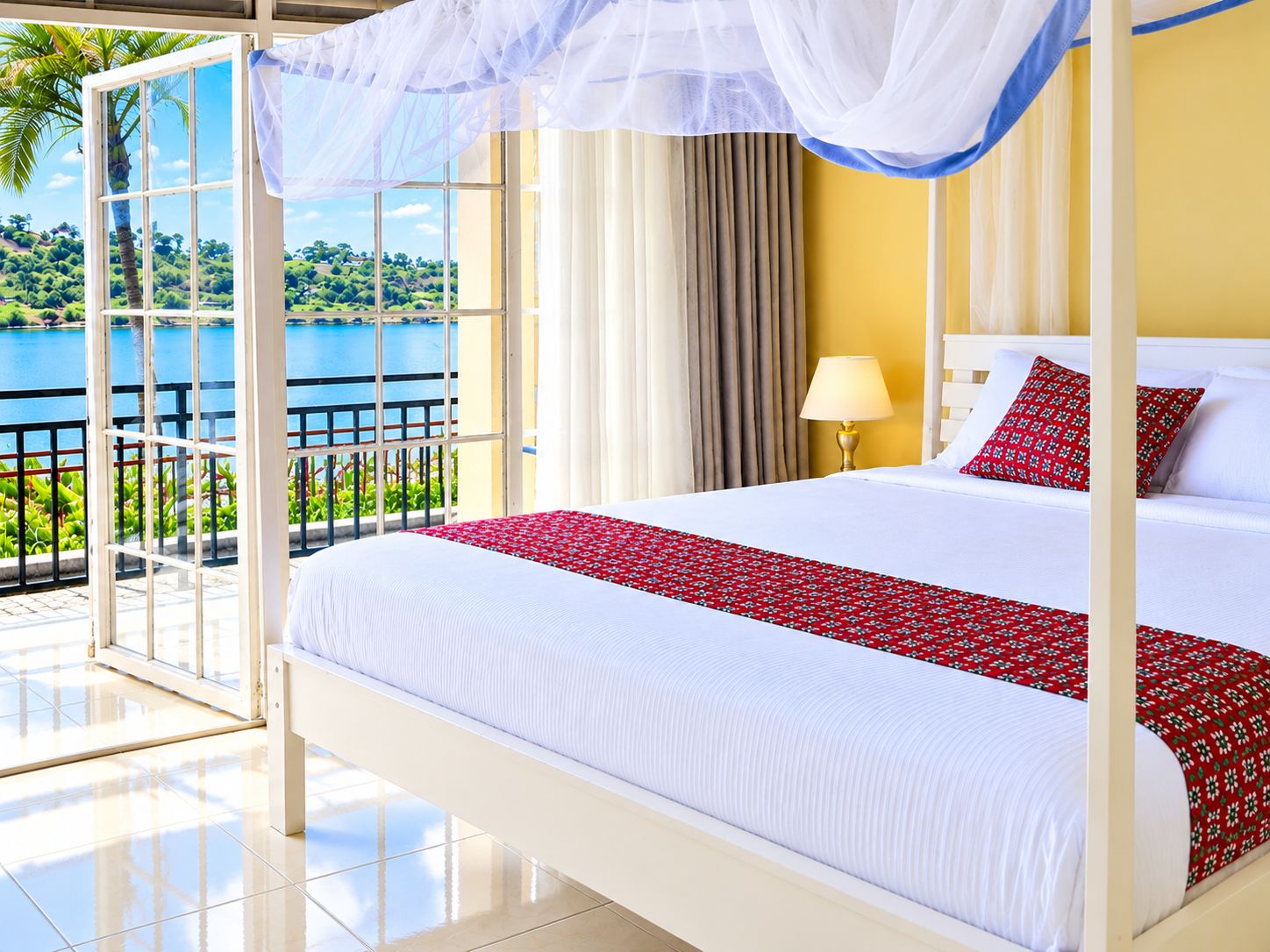A four-poster bed with a white and red bedspread, a nightstand with a lamp, and a view of a lake through a glass door.