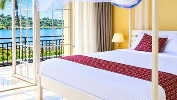 A four-poster bed with a white and red bedspread, a nightstand with a lamp, and a view of a lake through a glass door.