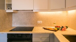 Eine Küche mit schwarchem Induktionskochfeld, einem Backofen und weißen Schränken. Auf der Arbeitsfläche steht eine Obstschale mit Erdbeeren und einer Zitrone.