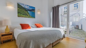 Une chambre à coucher avec un lit, deux oreillers, une table de chevet avec une lampe, une grande fenêtre avec des rideaux et un tableau représentant un paysage.