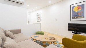 Una sala de estar moderna con un sofá color beige, un reposapiés verde, una mesa de centro redonda de madera y un sillón amarillo. Un televisor de pantalla plana está montado en la pared, y arriba hay un cuadro enmarcado de Portugal.