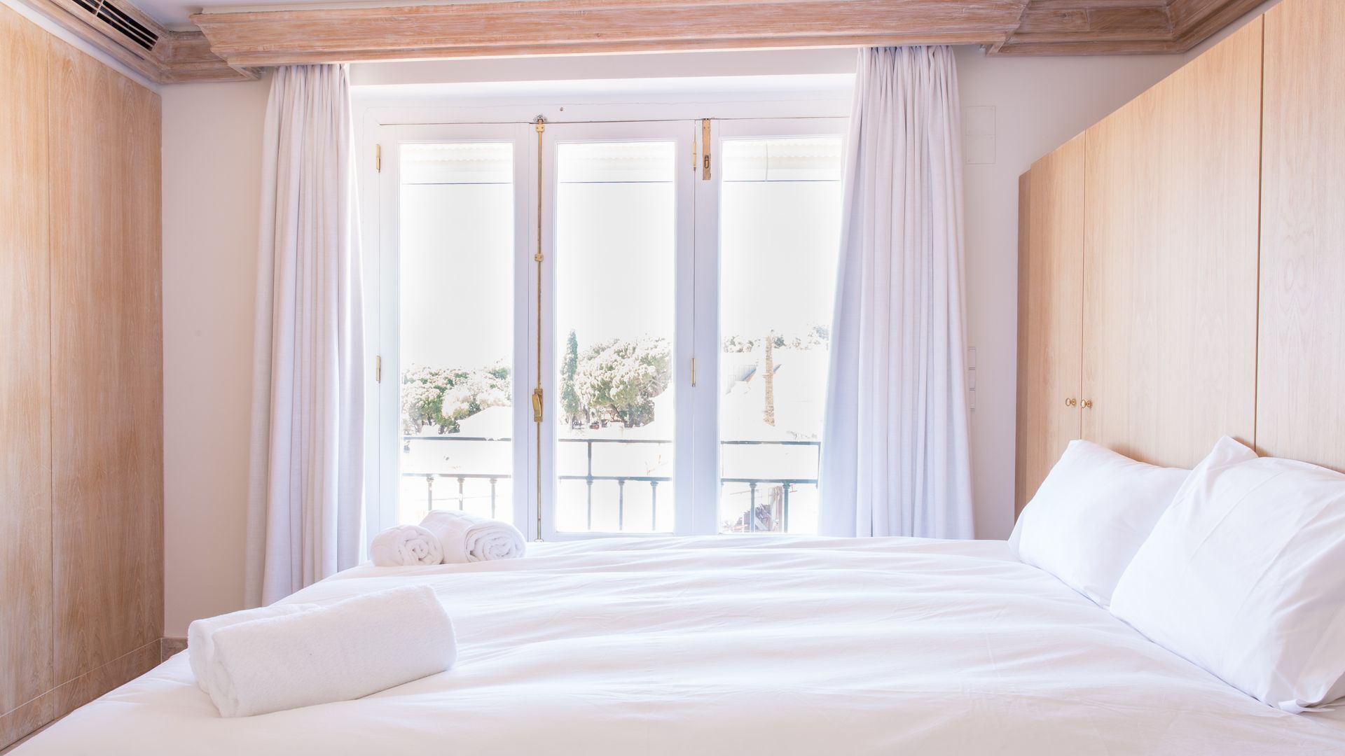 A hotel room with a large bed, white linens, a wooden headboard, and a balcony with curtains.