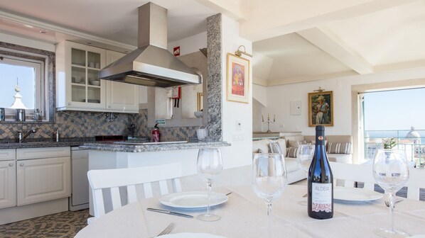 Cocina moderna con gabinetes blancos, encimera de granito y extractor de acero inoxidable. Hay un comedor con una mesa preparada para dos personas, que incluye copas de vino y una botella.