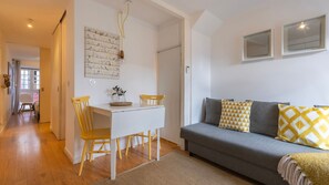 Ein modernes Wohnzimmer mit einem grauen Sofa, gelb-weißen Kissen mit Muster, einem weißen Esstisch und zwei gelben Stühlen sowie einer Wandleuchte.
