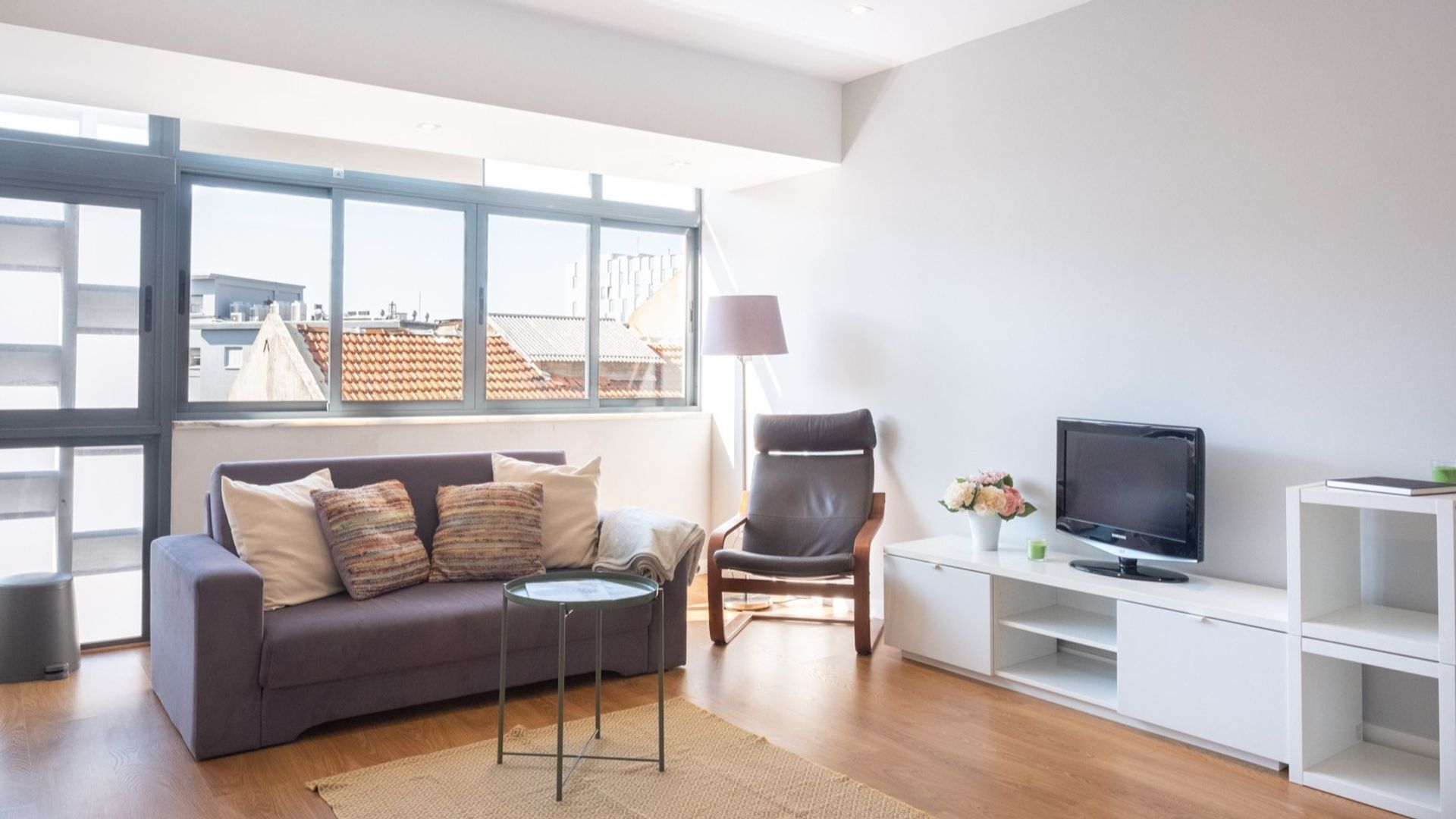 En moderne stue med en grå sofa, et lille rundt bord, en læderlænestol, et fjernsyn på et hvidt skab og et stort vindue med udsigt til bygninger.