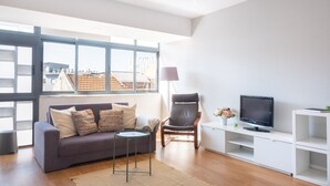 Un salon moderne avec un canapé gris, une petite table ronde, un fauteuil en cuir, un téléviseur posé sur un meuble blanc et une grande fenêtre donnant sur des immeubles.