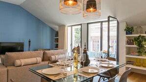 Un soggiorno moderno con un tavolo da pranzo in vetro apparecchiato per sei persone, un divano con cuscini decorati e una libreria piena di libri e oggetti ornamentali.