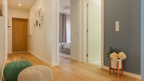 Un corridoio con pavimento in legno, un pouf beige e verde lavorato a maglia, un punto luce a parete e una porta che dà su una camera da letto.