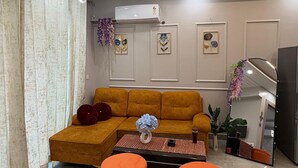 A modern living room with a sectional sofa, two ottomans, a coffee table, and decorative wall art. - A cozy and comfortable  1bhk apartment in Noida near Business Park and Mall (Noida)