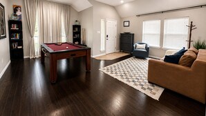 Een woonkamer met een pooltafel, een bank, een boekenkast en een deur die naar een andere kamer leidt.