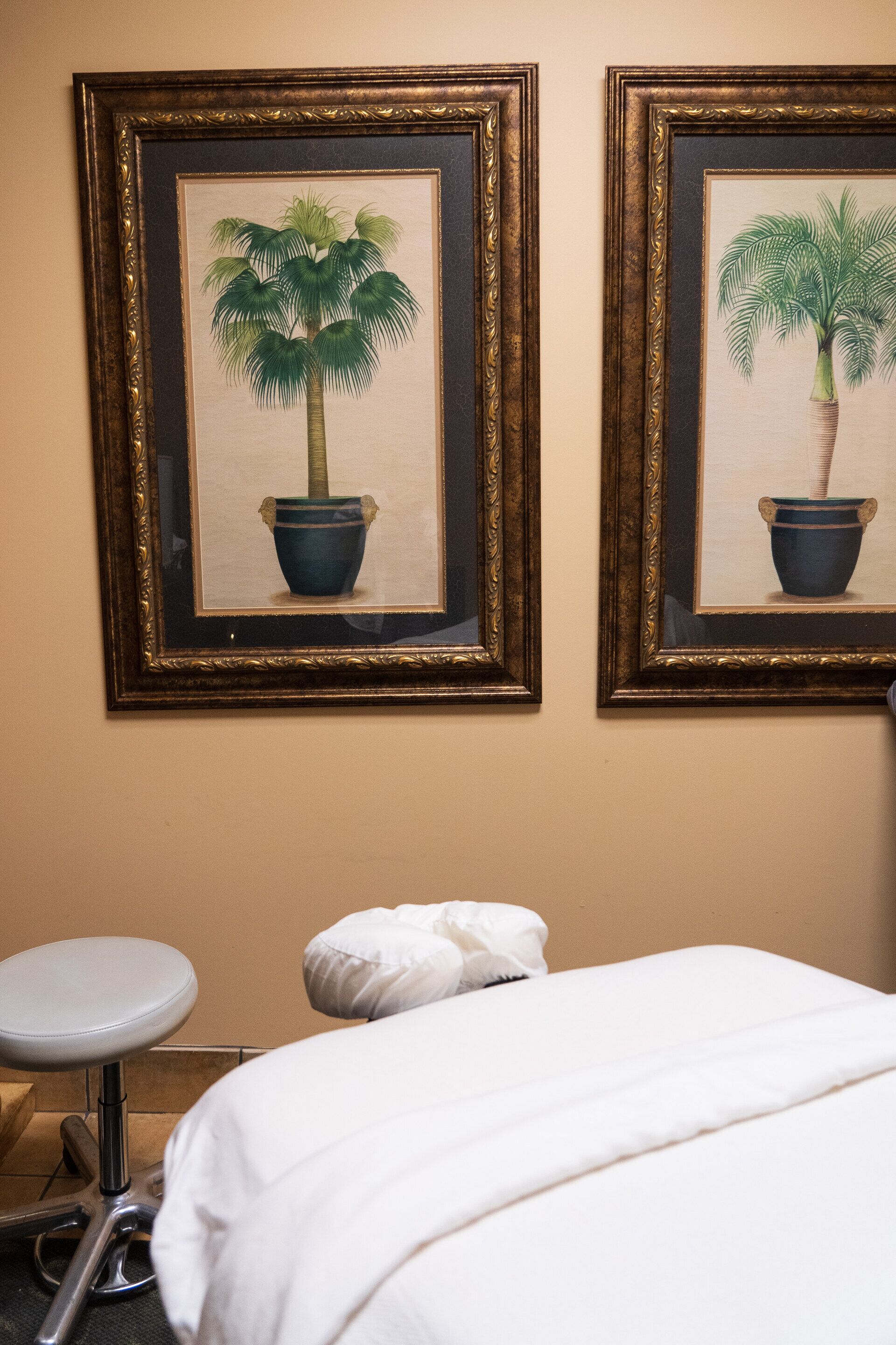 Habitación de hotel con cama, un taburete pequeño y dos cuadros enmarcados de palmeras.