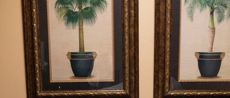 Habitación de hotel con cama, un taburete pequeño y dos cuadros enmarcados de palmeras.