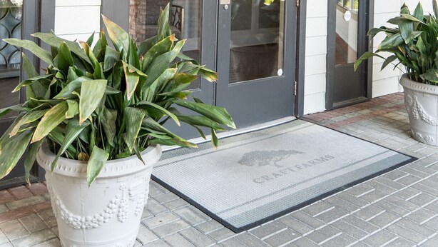L’entrée d’un immeuble, avec une porte sombre, une enseigne indiquant « FERME ARTISANALE » et deux plantes en pot placées sur les marches.