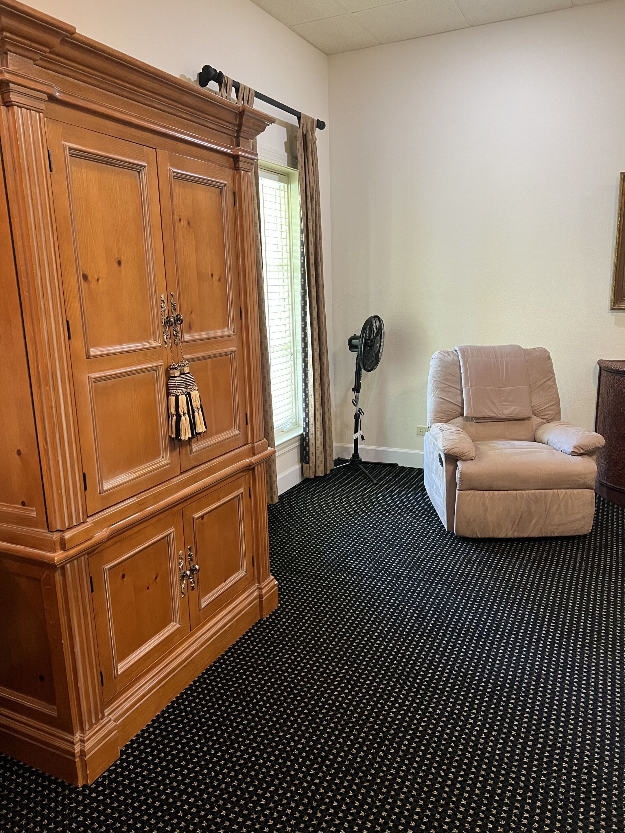 Ein Zimmer mit einem großen Holzschrank, einem beigen Sessel und einem Standventilator.