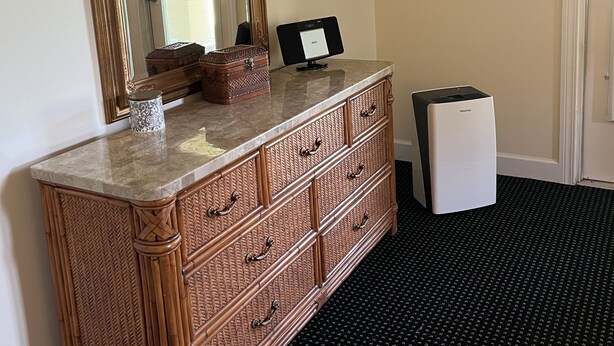 A room with a large mirror, a framed picture on the wall, a wooden dresser with drawers, and a trash bin. - The Villas at McCollough (Gulf Shores)