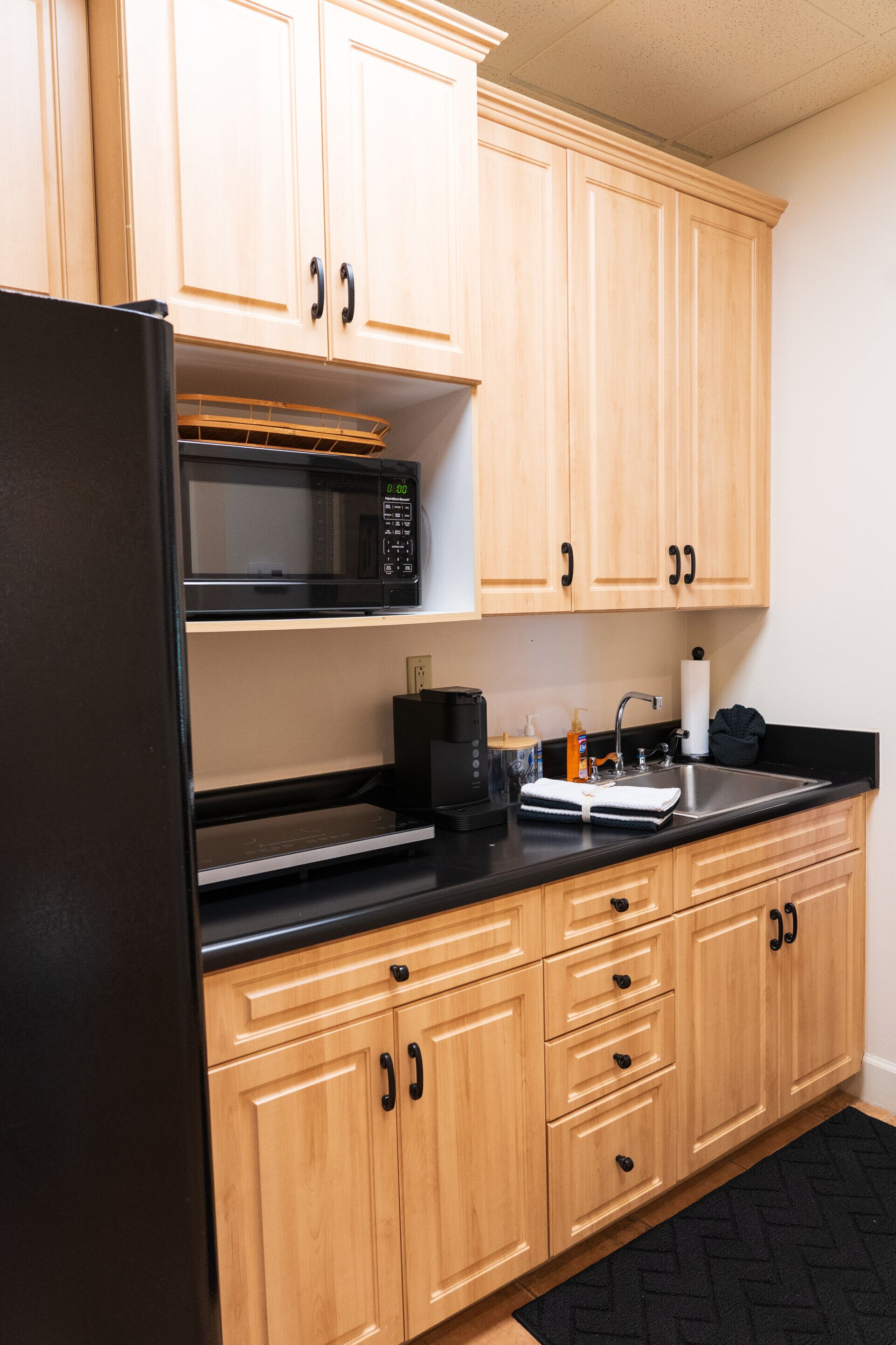 A kitchen with wooden cabinets, a black refrigerator, a microwave, a sink, and a coffee maker.