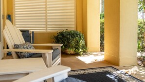 Une terrasse ensoleillée avec une chaise et une table blanches, une grande fenêtre avec des stores et une plante en pot.