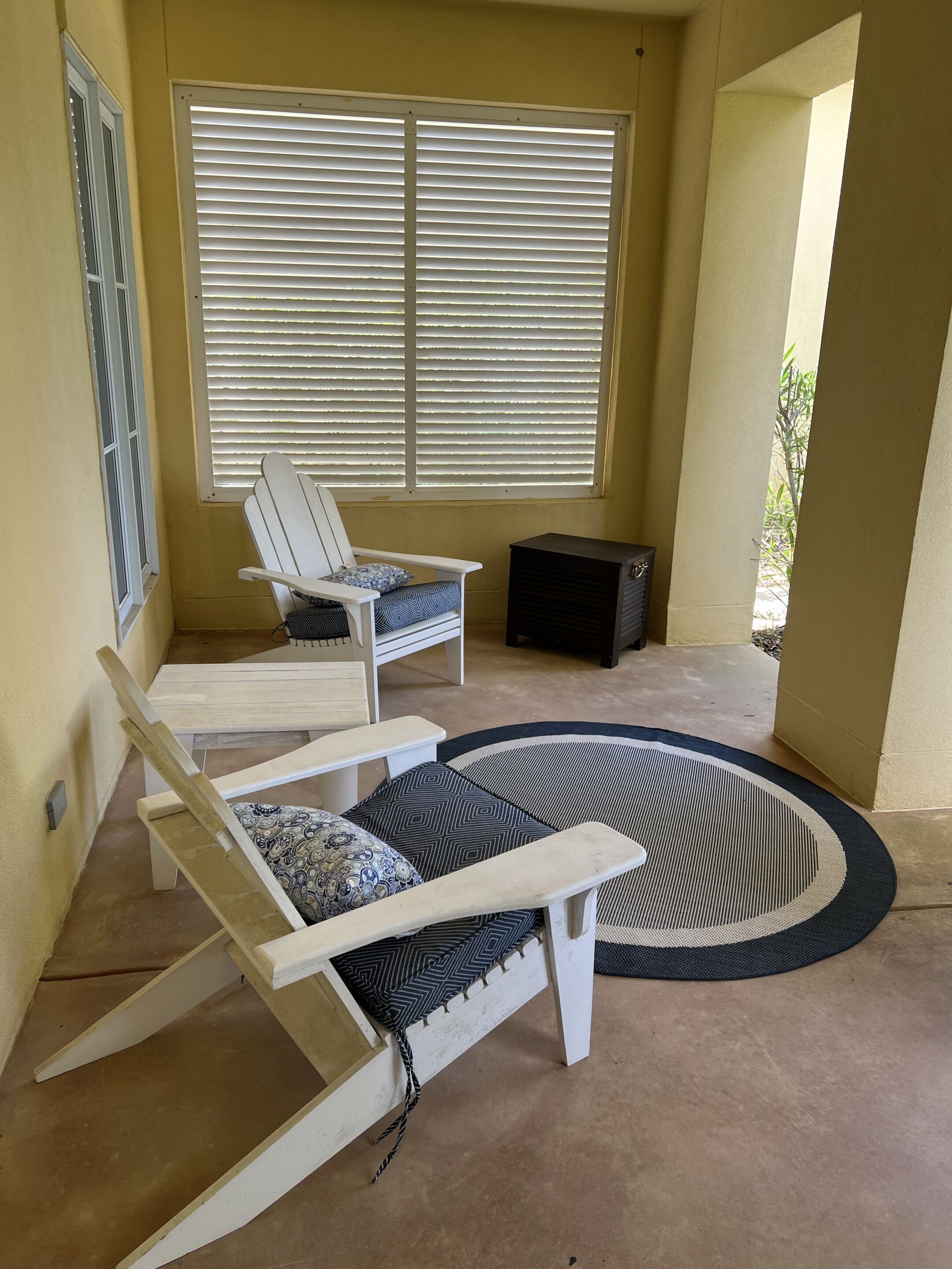 A sunlit room with a white chair and a white sofa with patterned cushions, a black heater, and a large window with blinds.