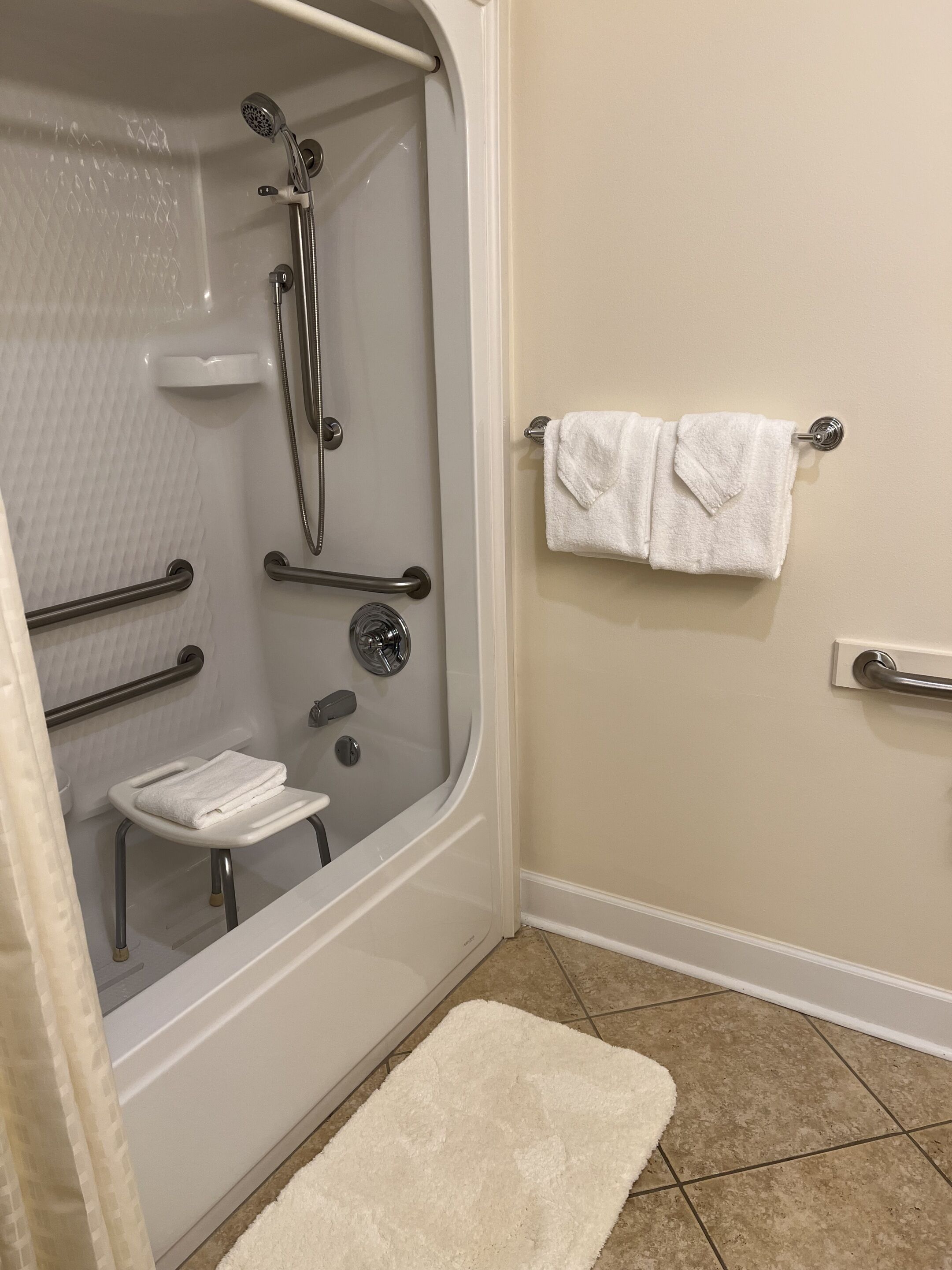 Un baño con ducha a ras de suelo, barras de apoyo, una toalla blanca sobre un perchero y un tapete de baño blanco.