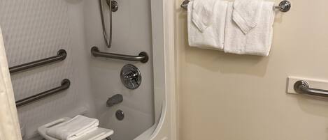 Un baño con ducha a ras de suelo, barras de apoyo, una toalla blanca sobre un perchero y un tapete de baño blanco.