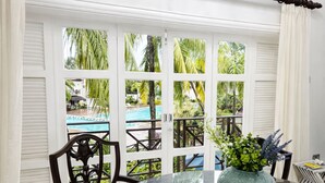 A dining area with a round table, chairs, a vase with flowers, and a view of a pool through a window with shutters. - Cap Cove 3BR Villa • Pool • 24/7 Security • Beach Access (Gros Islet)