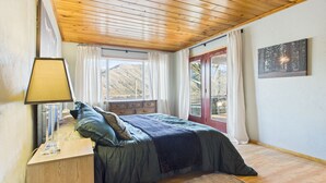Une chambre avec un plafond en bois, un lit recouvert d’une courtepointe bleue, une table de chevet avec une lampe, une fenêtre avec des rideaux et une porte donnant sur la vue des arbres.