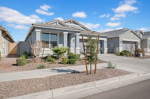 Une maison d’habitation à la façade grise, dotée d’un porche et d’un jardin aménagé avec de petits arbres et des arbustes.