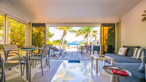 A modern living room with a glass door leading to a deck with outdoor furniture, a sofa, and a coffee table. - Cottage Beach House NBBC 1 – Beachfront Escape, Just Steps from the Sea (Baie Nettlé)