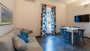 A living room with a sofa, a dining table with chairs, a TV mounted on the wall, and a window with patterned curtains. - AMARI APARTMENT DOWNTOWN PALERMO (Palermo)
