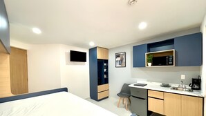 A compact hotel room with a bed, a desk, a chair, a microwave, a sink, and a small cabinet. - Living Alameda (Queretaro)