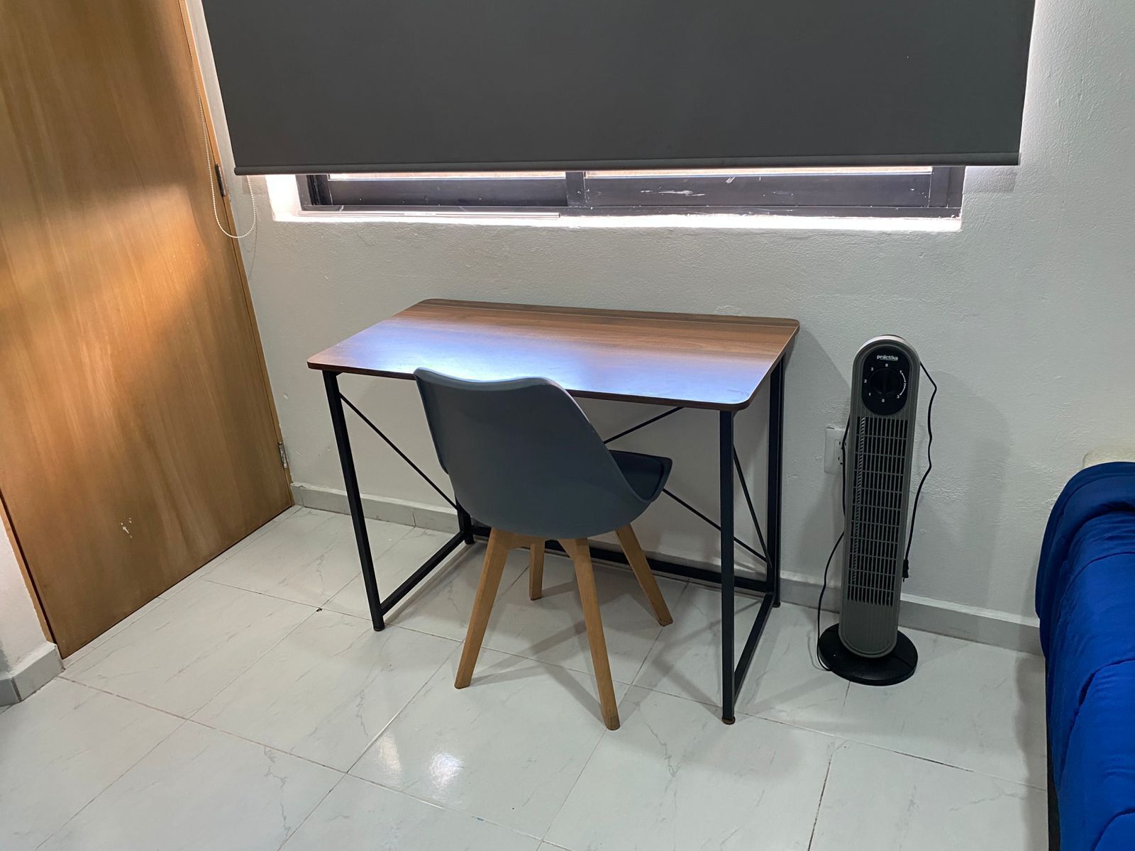 Una habitación pequeña con un escritorio, una silla, un ventilador de pie y un sofá azul.