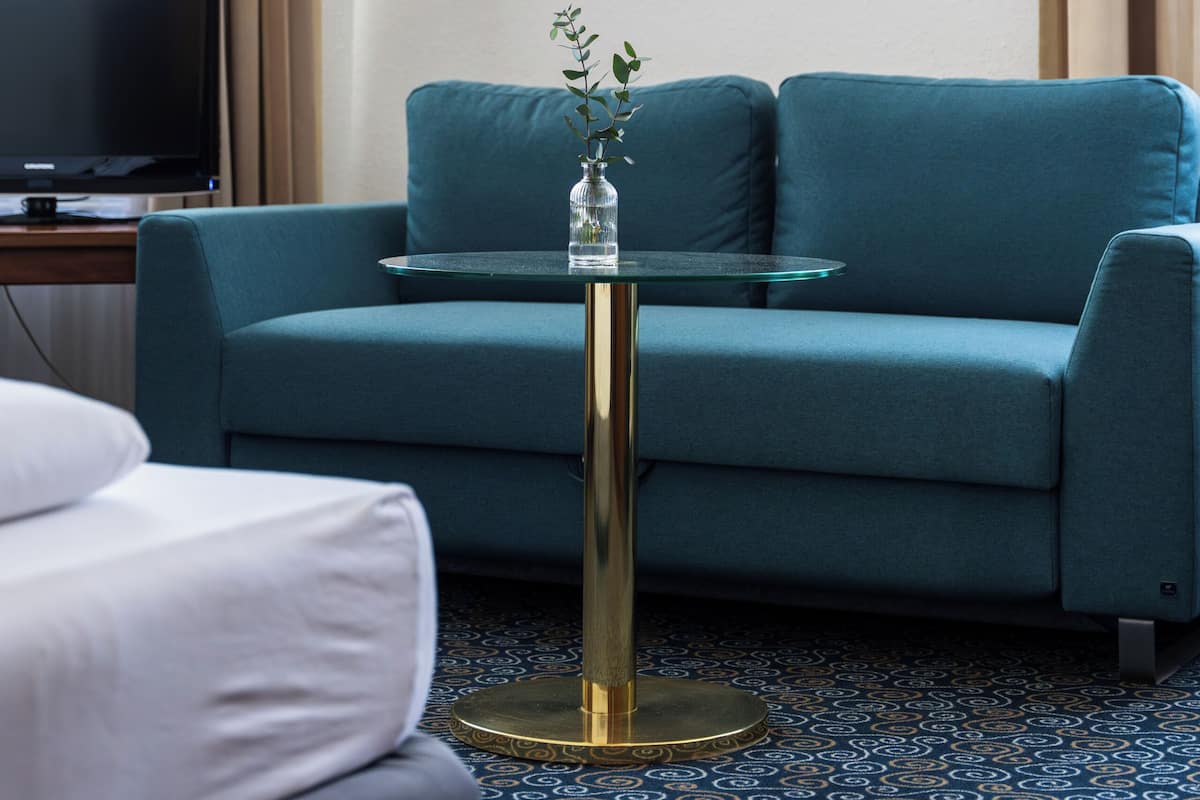 A hotel room with a teal sofa, a glass coffee table, a framed photograph on the wall, and a television on a wooden stand.