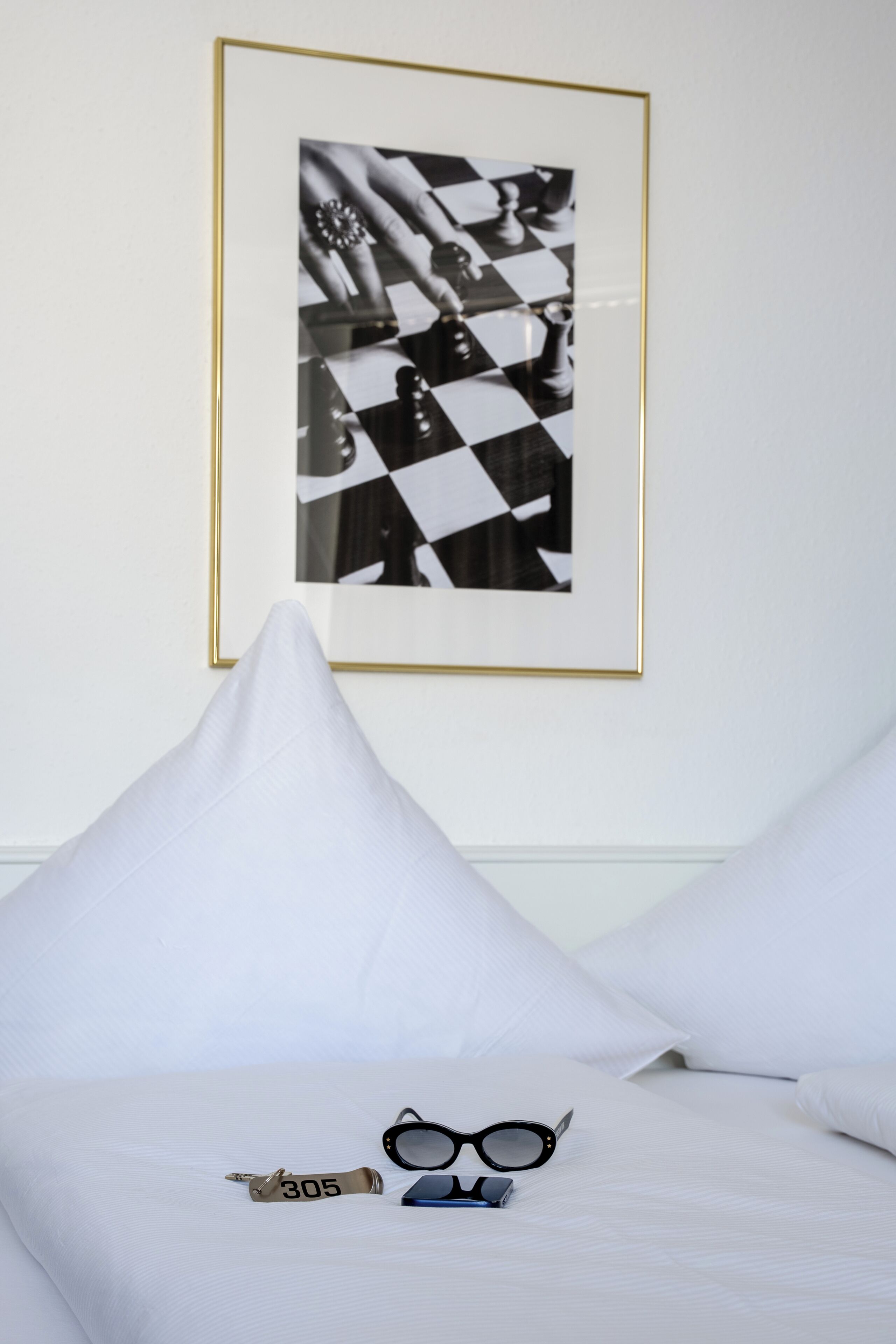 A neatly made bed with white bedding, a framed chessboard artwork on the wall, and a pair of glasses and a pen on the bed.