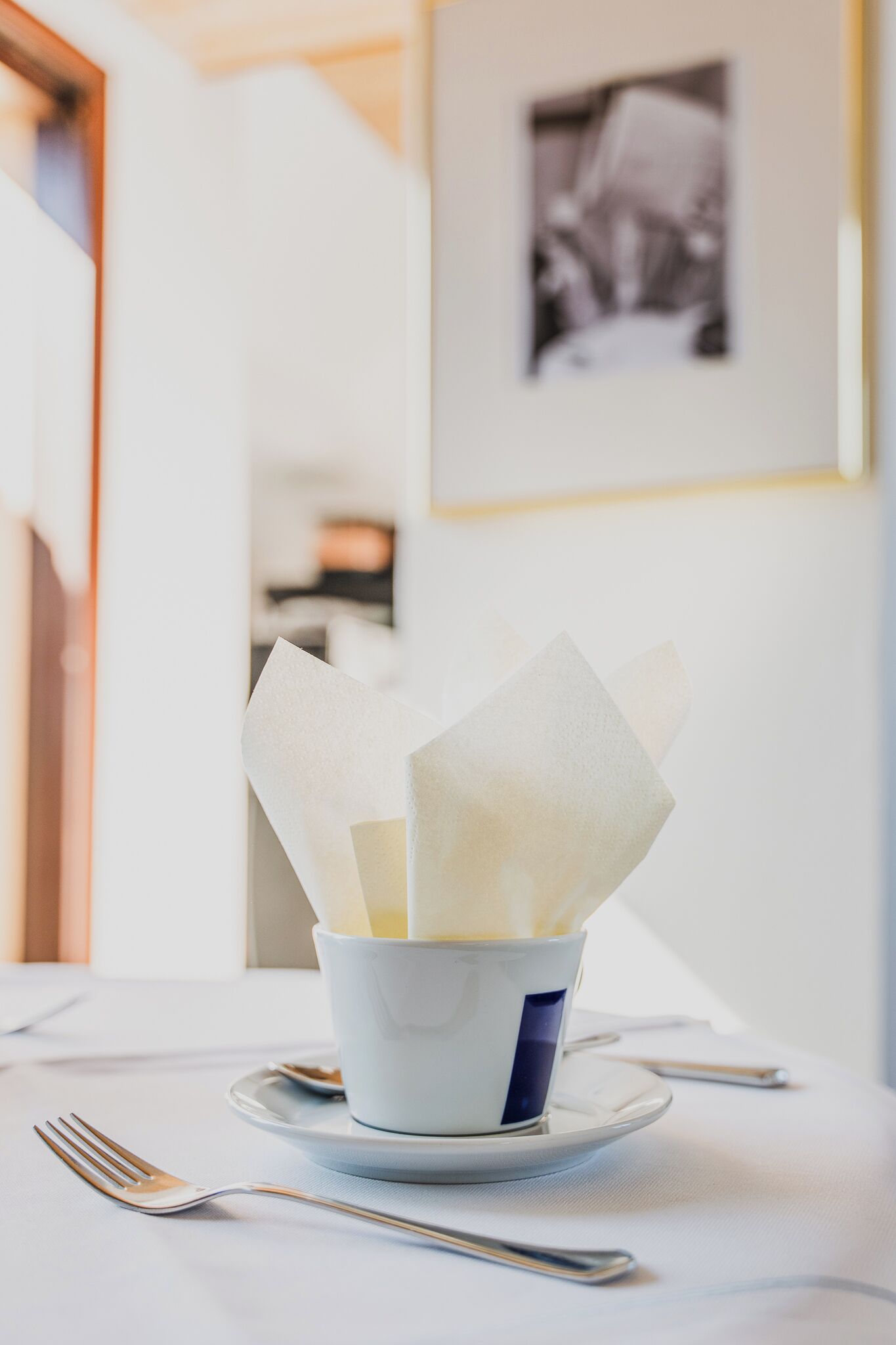 A cup of white food on a white table with a fork and spoon beside it, a framed picture on the wall, and a door in the background.