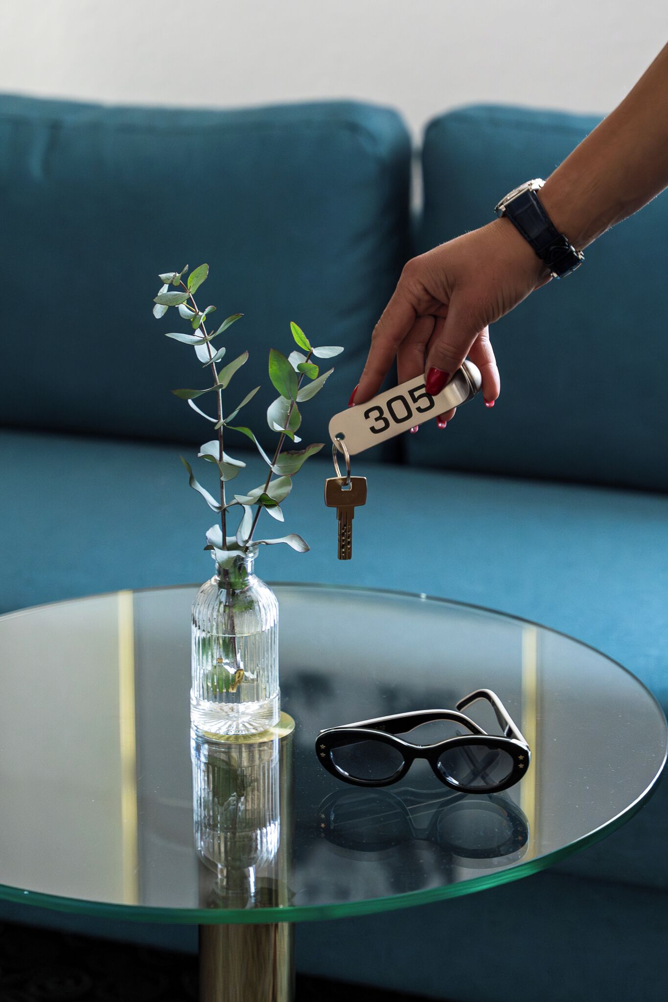 Una mano sosteniendo una llave con el número 305, un florero de vidrio con una planta y un par de gafas sobre una mesa redonda de vidrio.