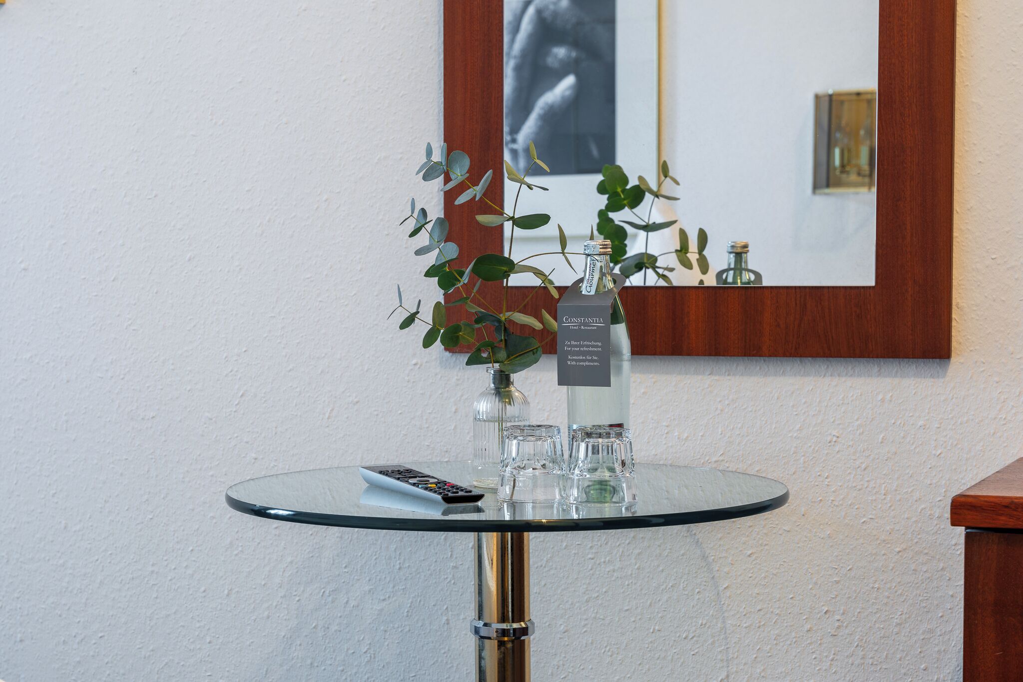 Una mesa de vidrio con un control remoto, una botella de Constantia y un florero con hojas verdes.