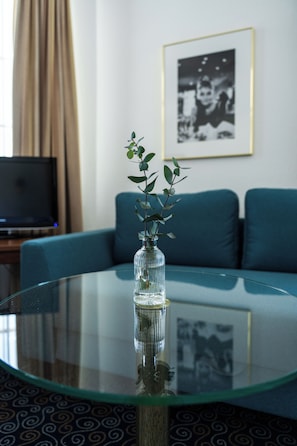 A glass table with a clear vase containing green leaves, a blue sofa, and a framed picture on the wall.