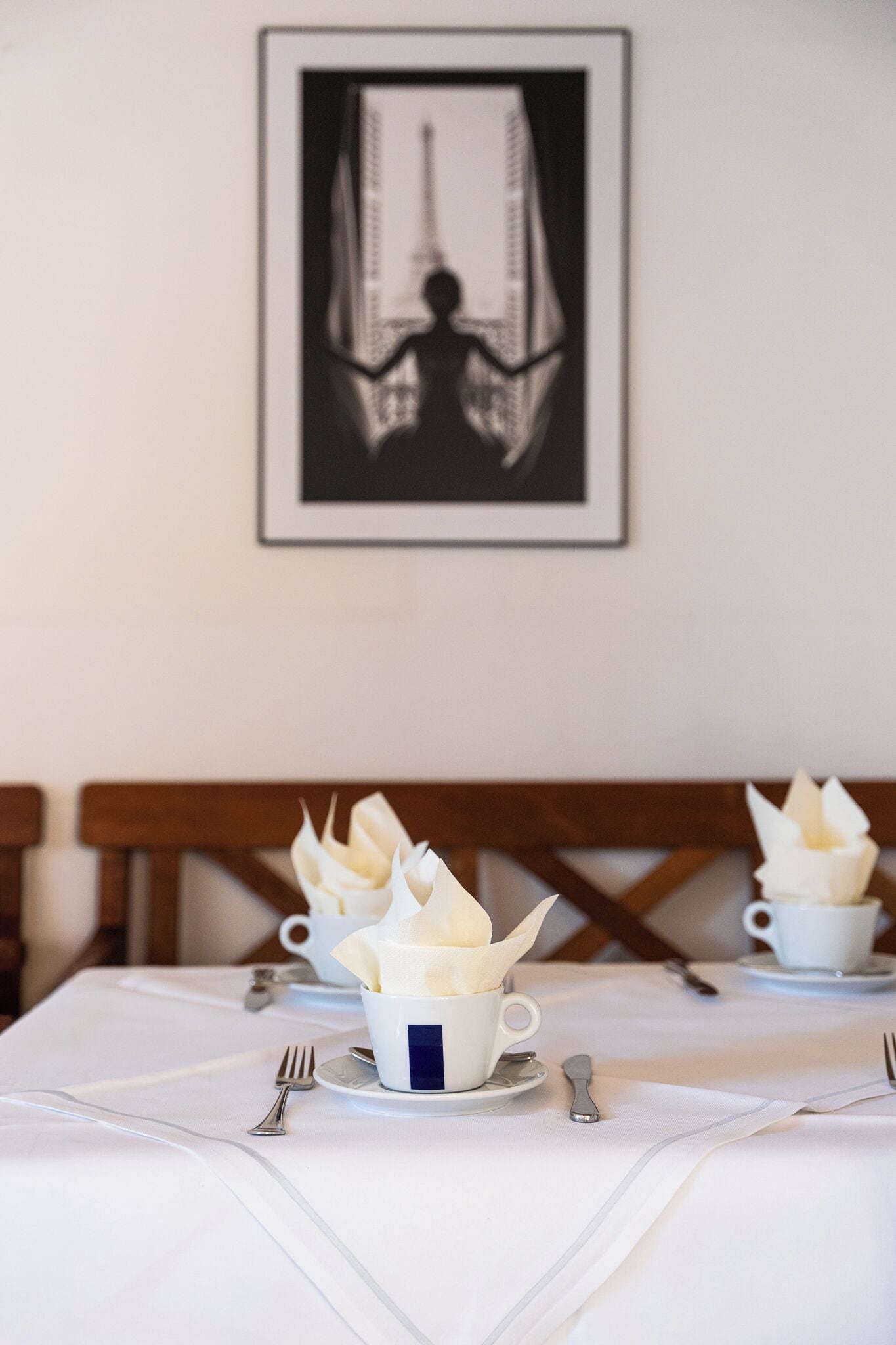 Una mesa de comedor con mantel blanco, tazas blancas con servilletas y cubiertos. Un cuadro enmarcado de una persona está en la pared detrás de la mesa.