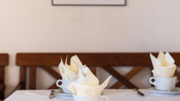 Una mesa de comedor con mantel blanco, tazas blancas con servilletas y cubiertos. Un cuadro enmarcado de una persona está en la pared detrás de la mesa.