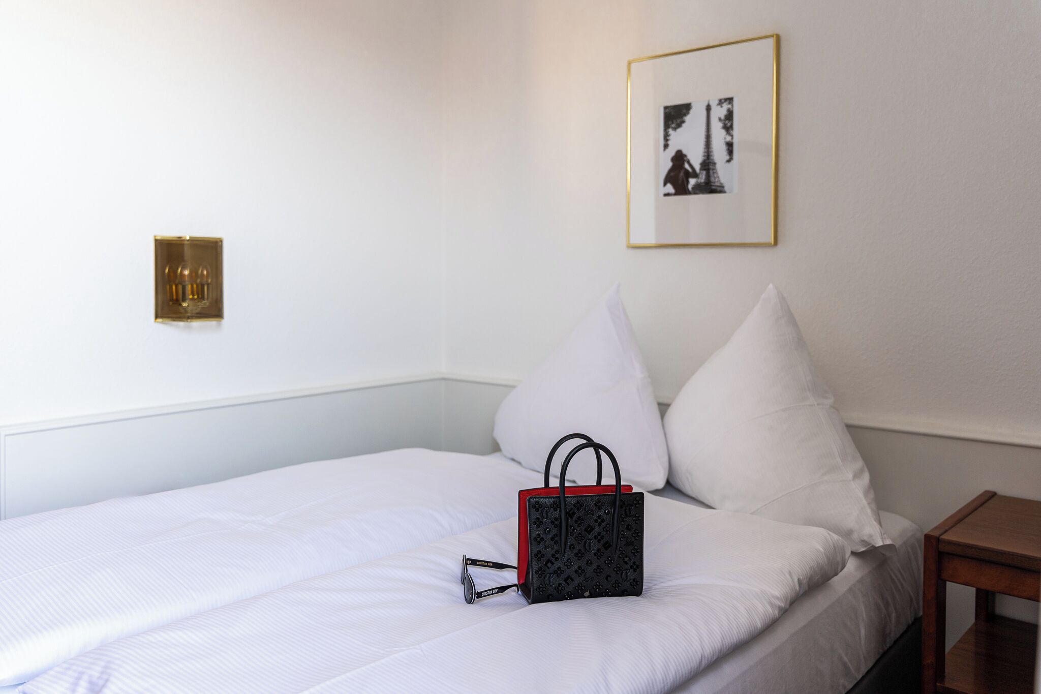 A neatly made bed with white linens, a black and red bag, and a framed picture on the wall.