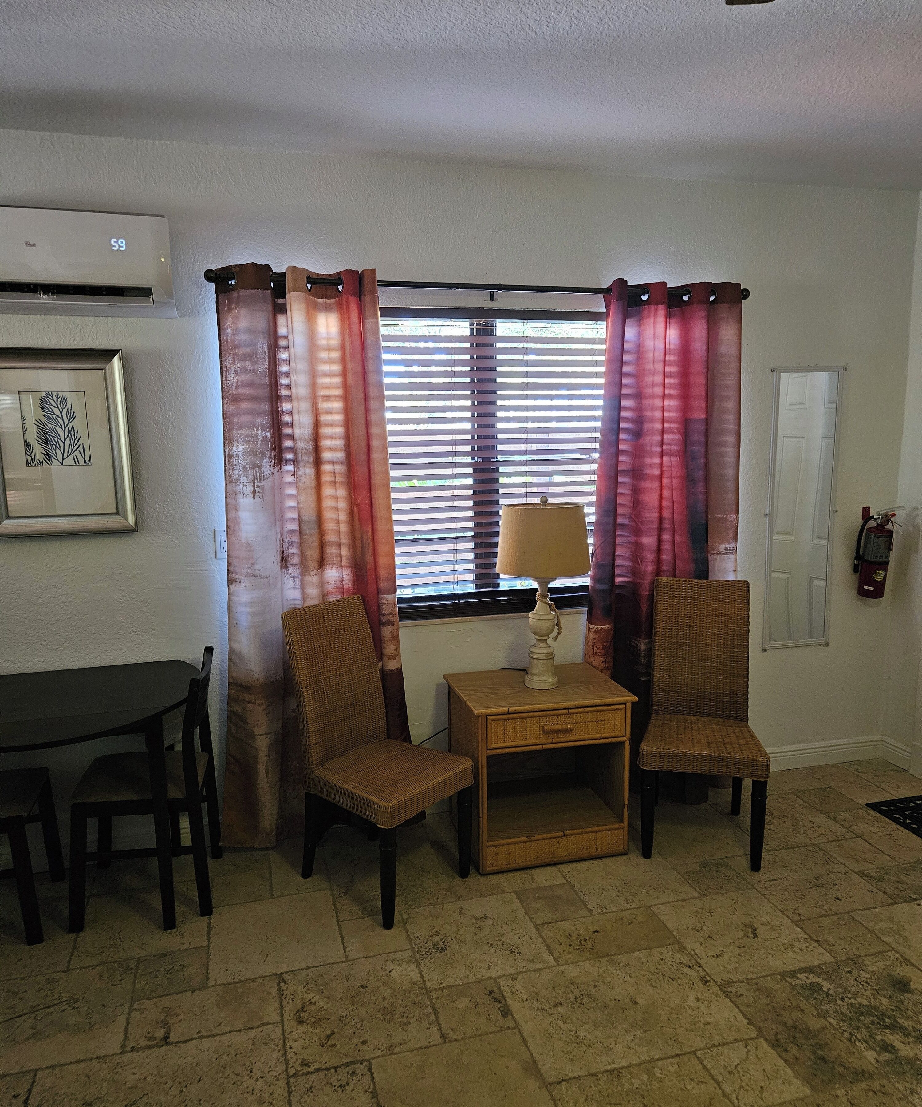 A living room with a dining table, two wicker chairs, a small wooden side table with a lamp, and a window with blinds and curtains.