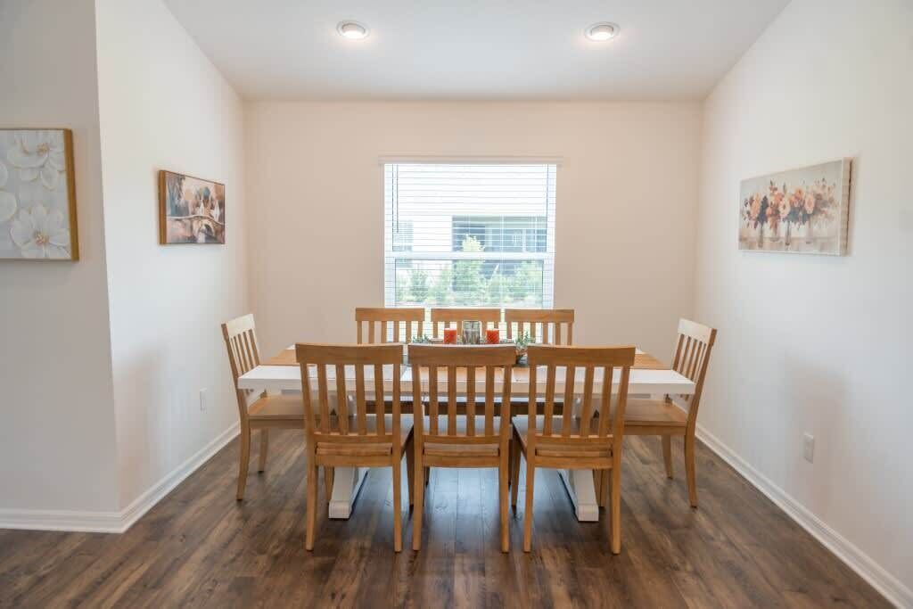 A dining area with a round table and six wooden chairs, a large window with blinds, and two framed pictures on the walls.