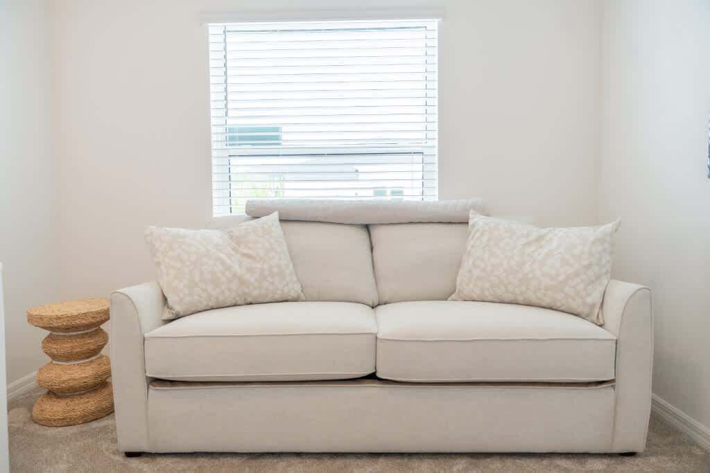 A white sofa with two patterned pillows, a woven side table, and a window with blinds.