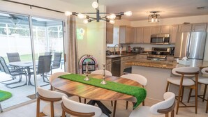 Une cuisine moderne dotée d’appareils électroménagers en acier inoxydable, un coin repas avec une table en bois et des chaises blanches, et une vue sur l’extérieur grâce à de grandes portes vitrées.